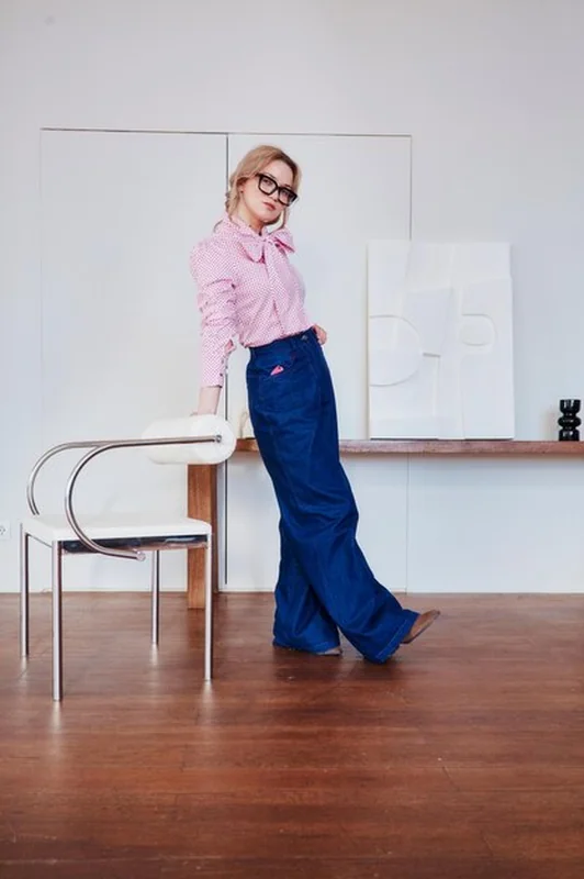 Stylowa kobieta w różowej koszuli i niebieskich dżinsach opierająca się o krzesło