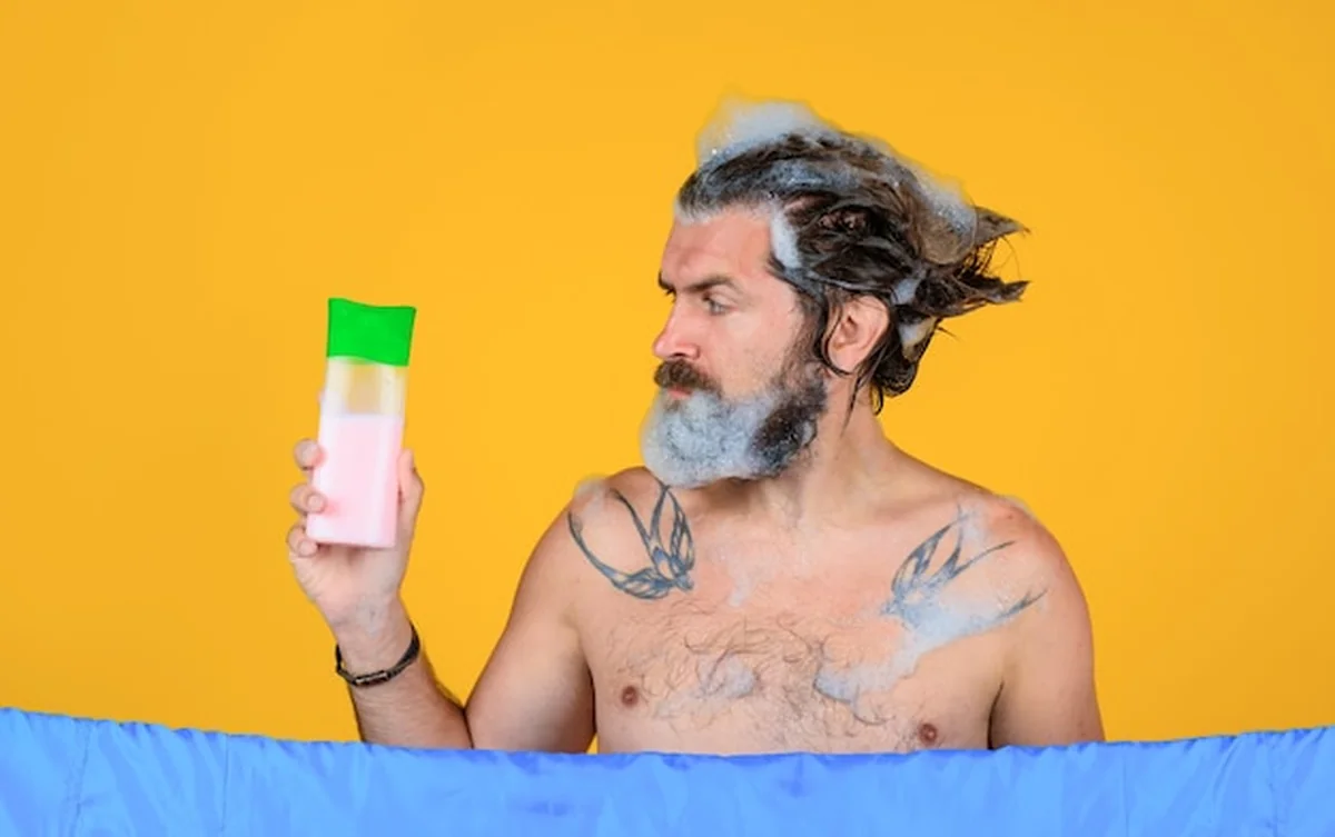 Mężczyzna myjący włosy szamponem pod prysznicem z pianką na głowie i butelką szamponu