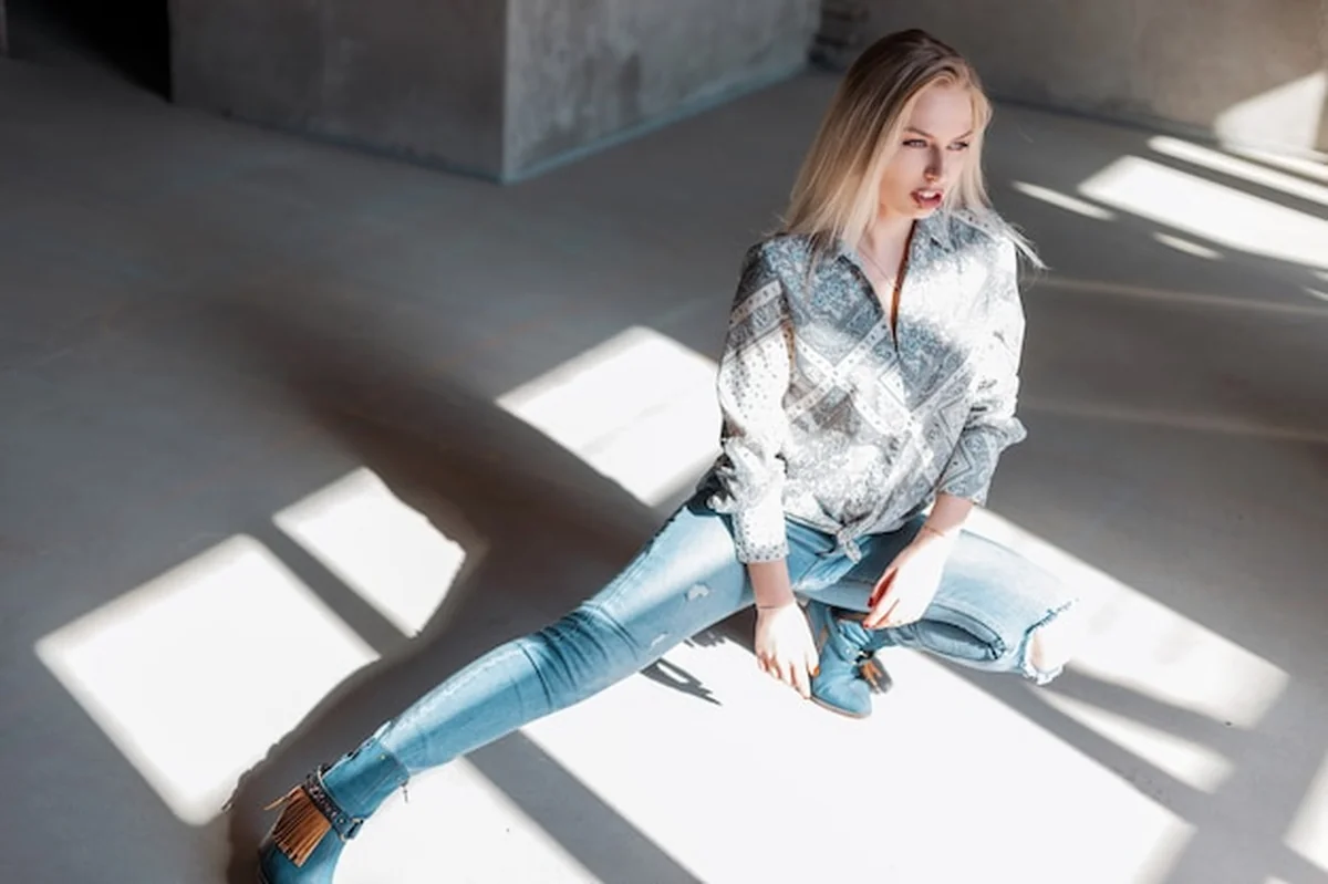 Młoda kobieta w szarych dżinsach i kowbojskich butach, pozująca w koszuli vintage w pomieszczeniu
